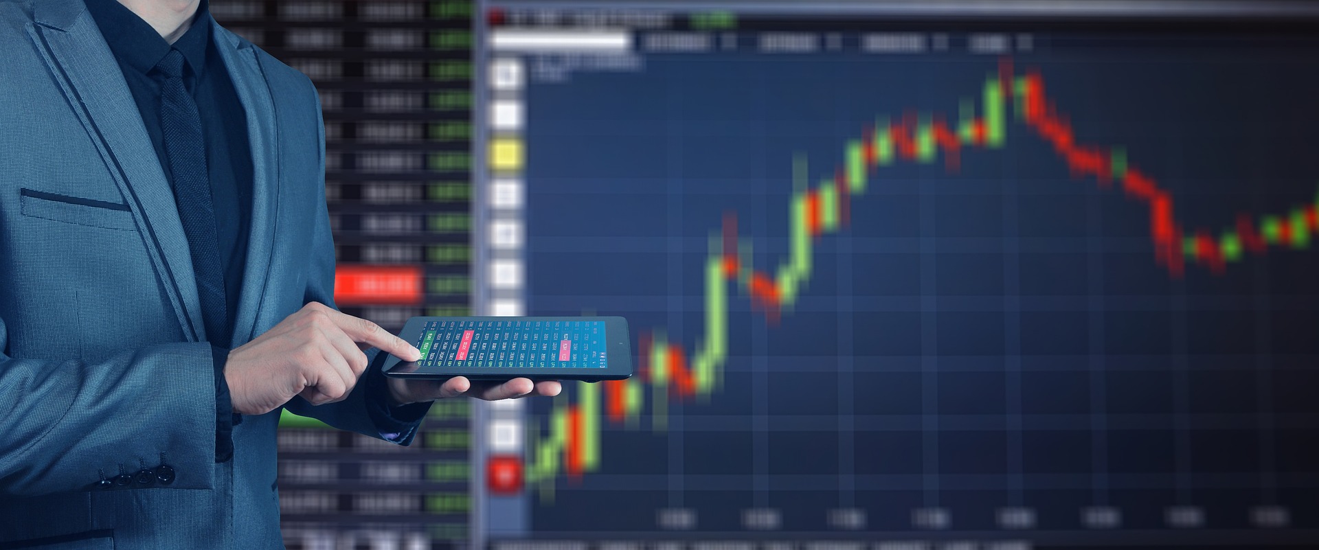 Une personne se tient avec une tablette devant un grand écran affichant l'évolution de la bourse